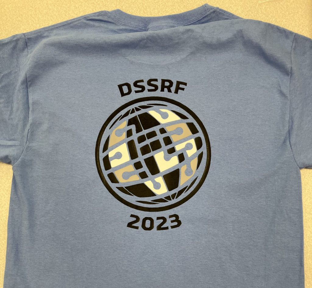 A medium blue shirt with a globe-like logo in blue, grey, and white that reads, "DSSRF 2023"