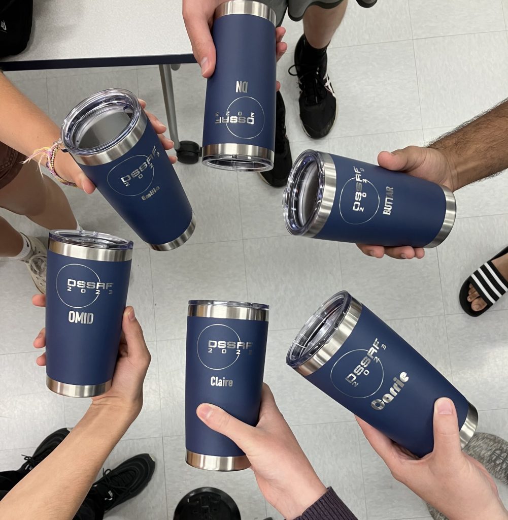 Six blue travel thermoses that have been laser engraved to say "DSSRF 2023," held in a circle by each member of the group